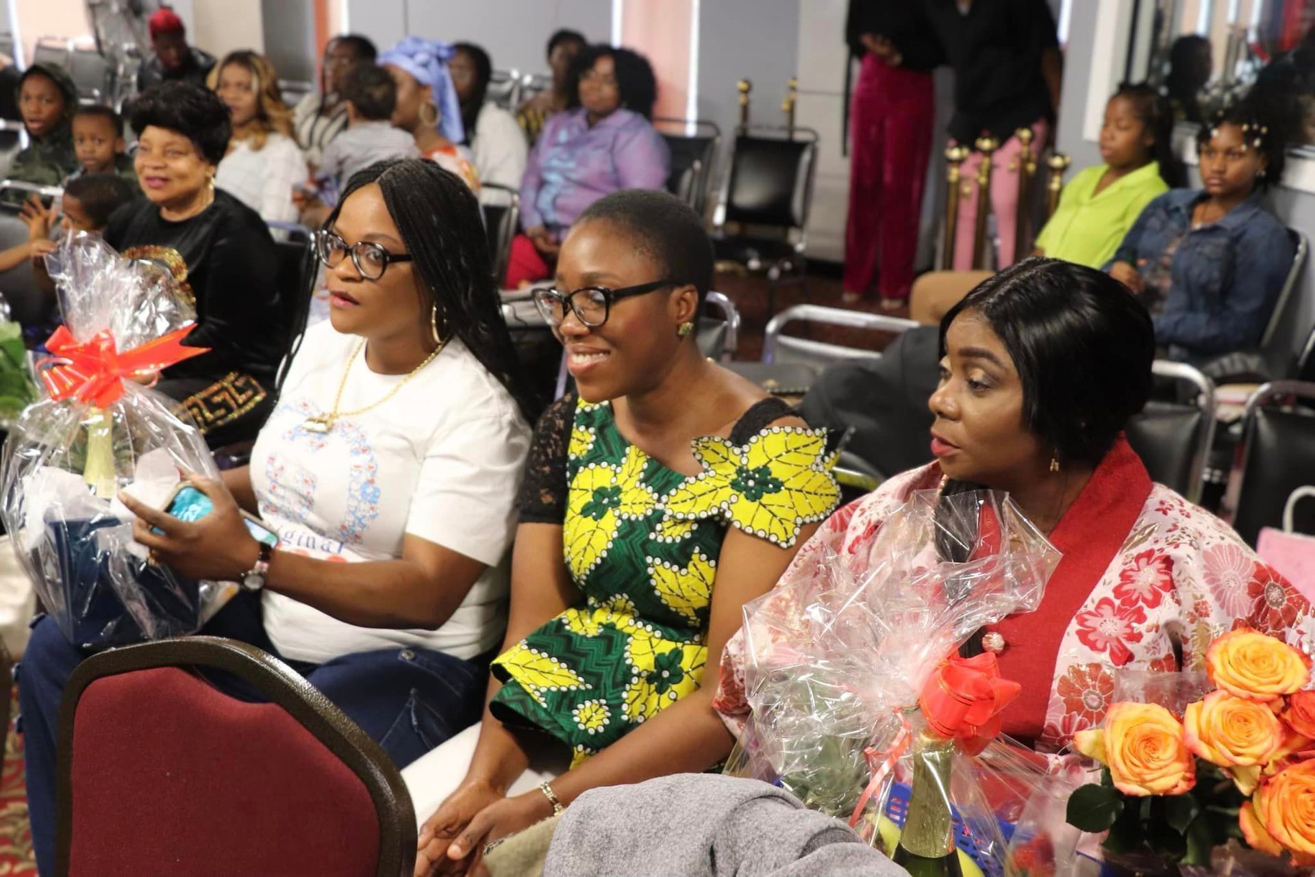 Group of women smiling, holding gifts, seated at an event. Brightly colored clothing and flowers are visible.