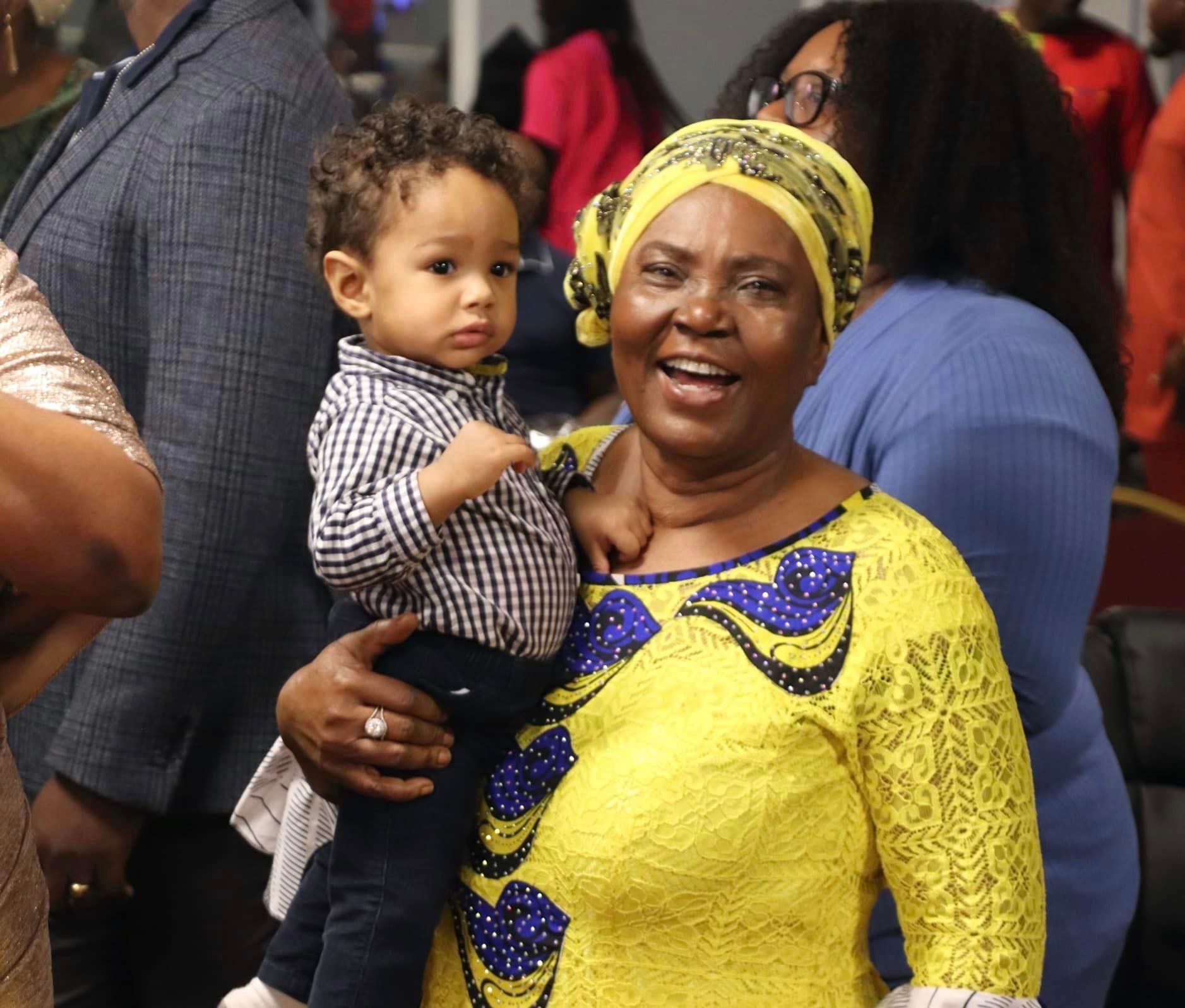 Smiling woman in yellow dress holding a young boy with curly hair; they're in a crowded indoor setting.