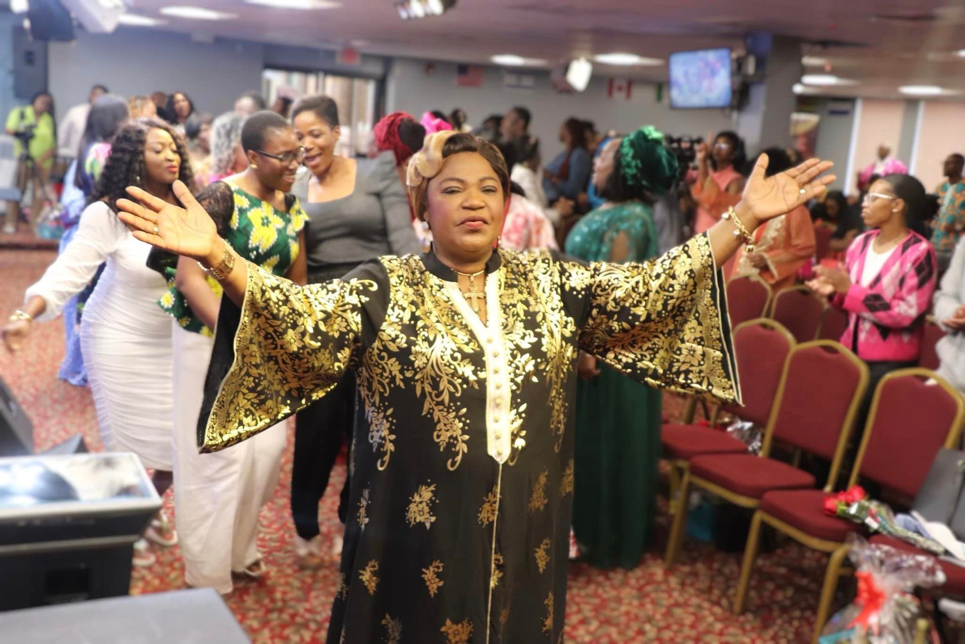 Woman in gold-trimmed black robe with arms outstretched, leading worship in a church. People in background.
