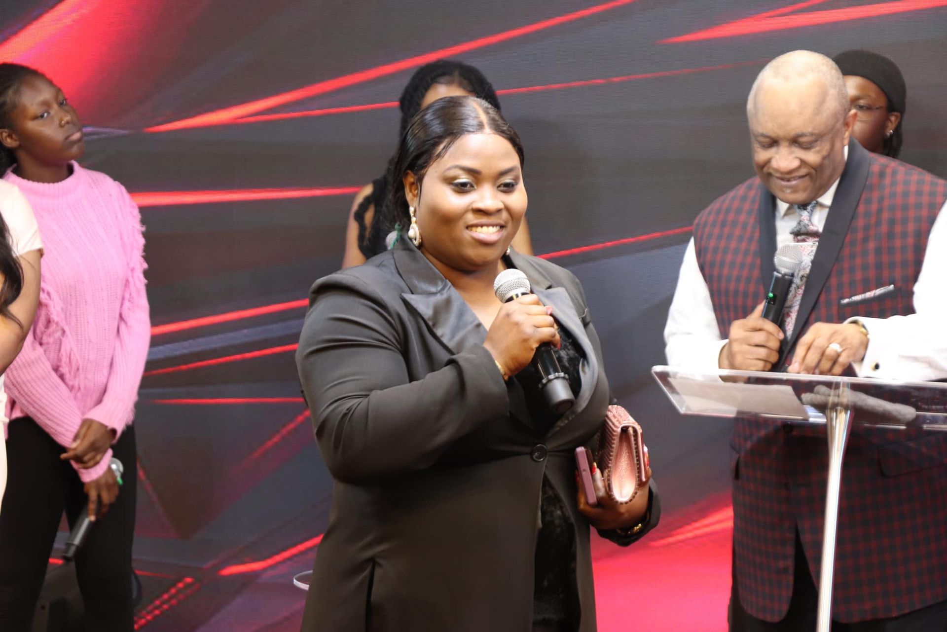 Woman in black suit speaks into a microphone, smiling, beside an older man at a podium. Others stand in the background.