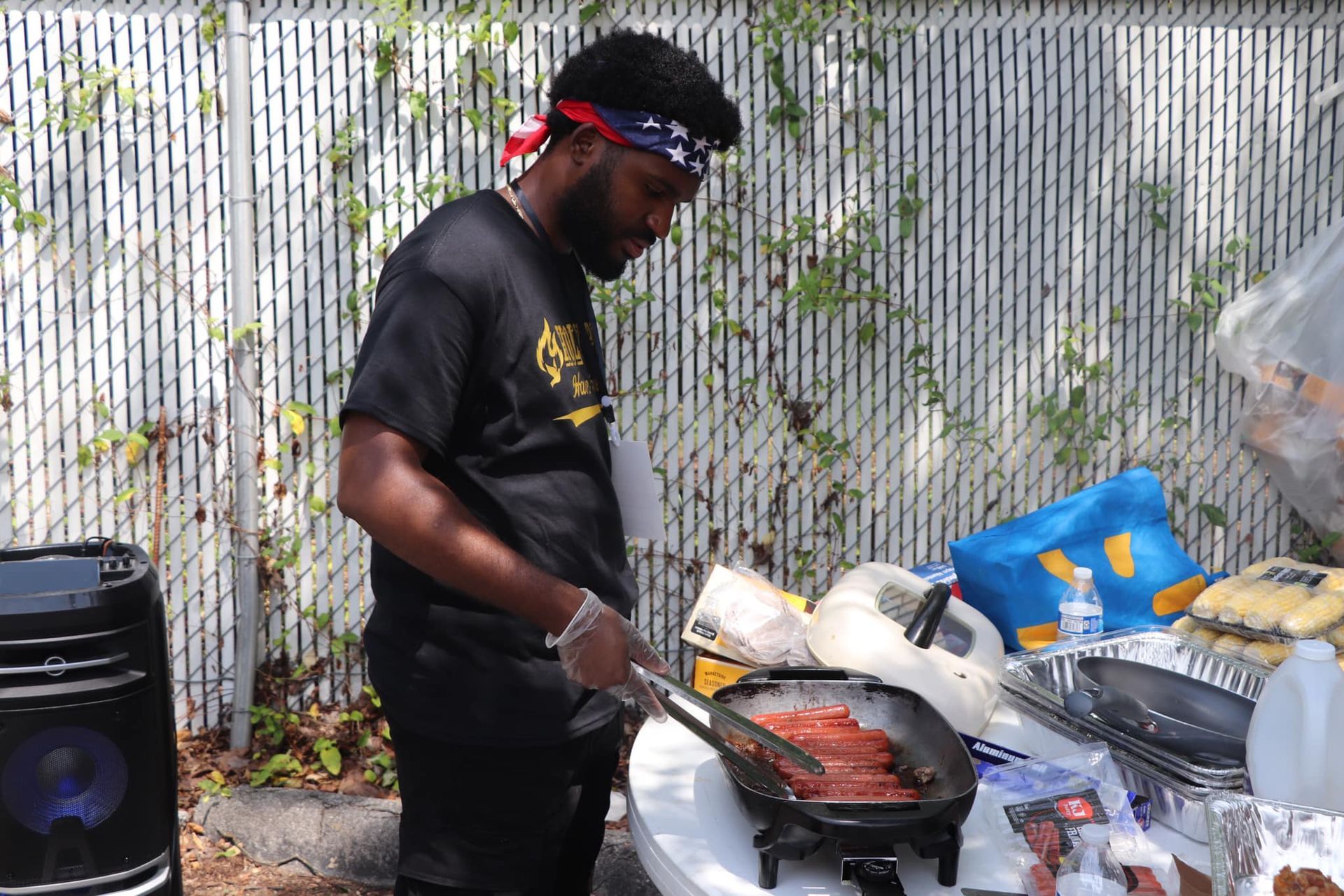 Man grilling food outdoors, wearing a patriotic headband, near a white fence.
