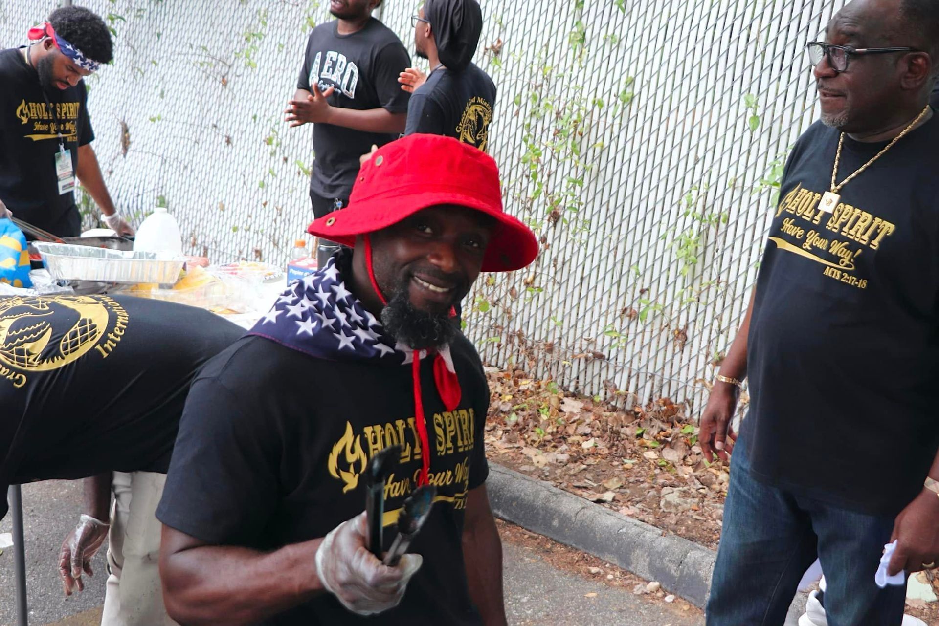Man in red hat smiles, holding item. Others wear black shirts, some cooking outdoors.
