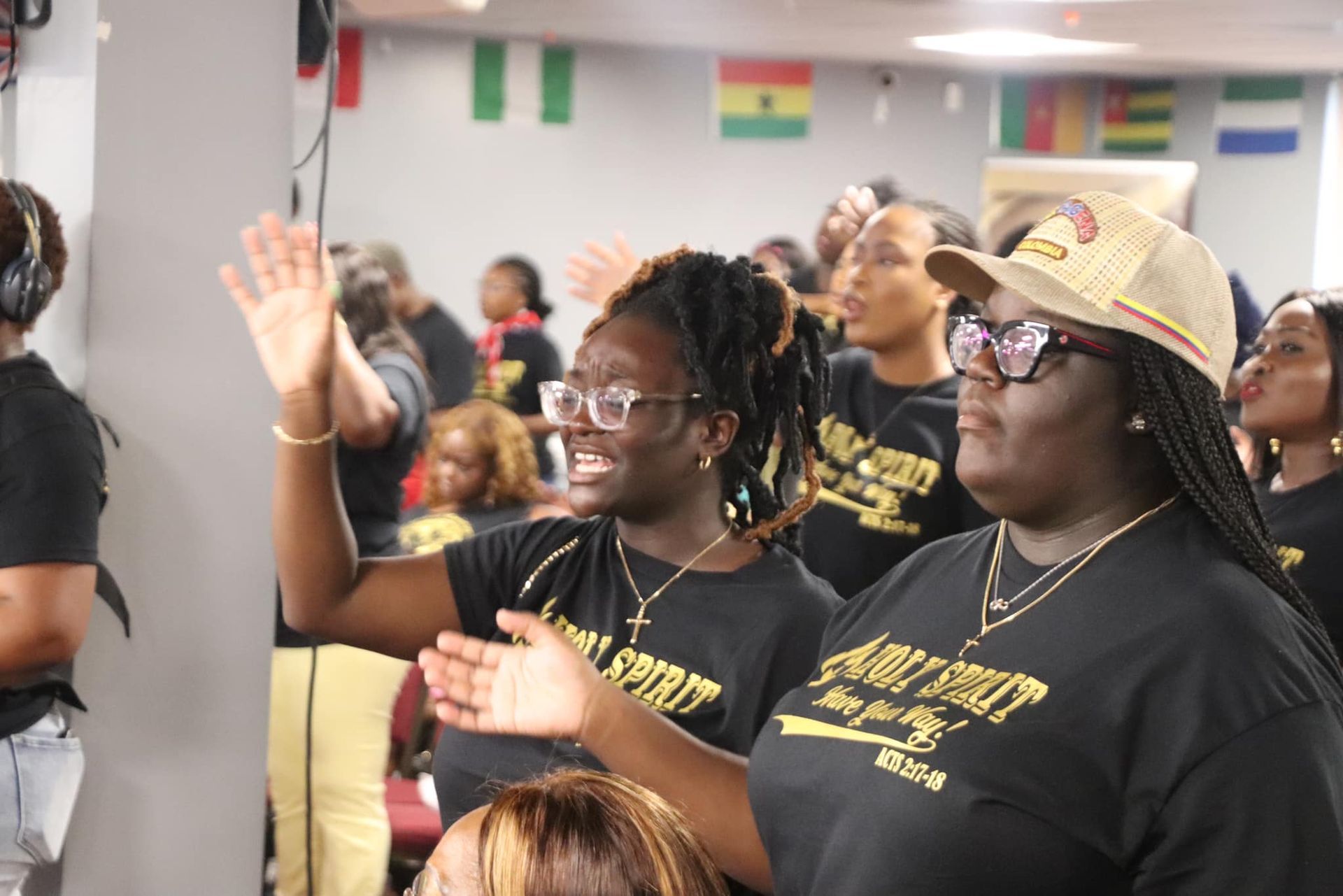 People in black shirts with gold lettering raising hands, smiling, in front of flags.