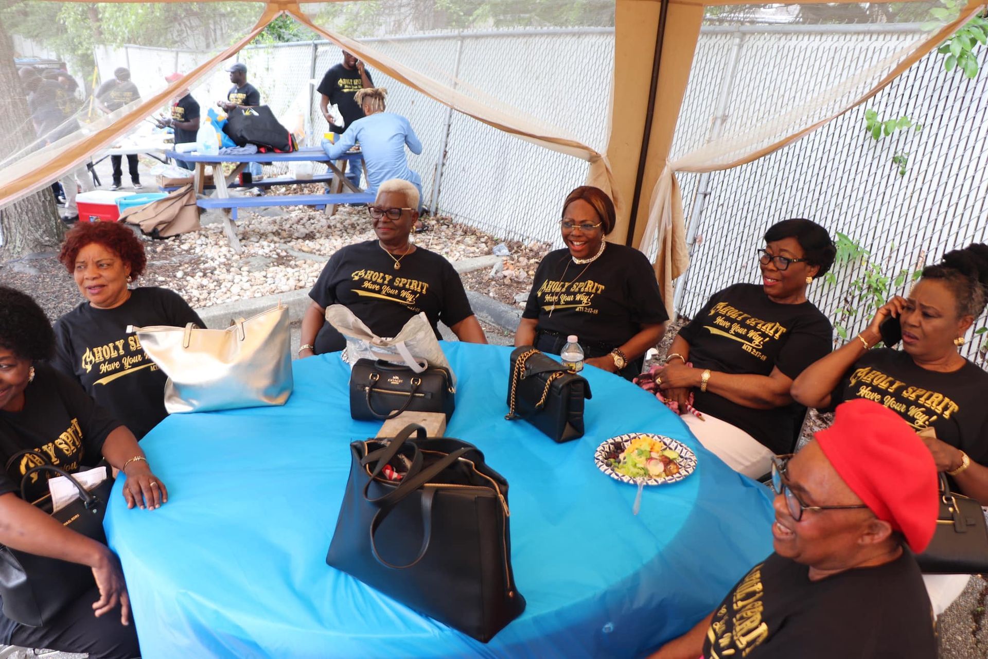 Group of women in black shirts, smiling at a round table, under a tent.