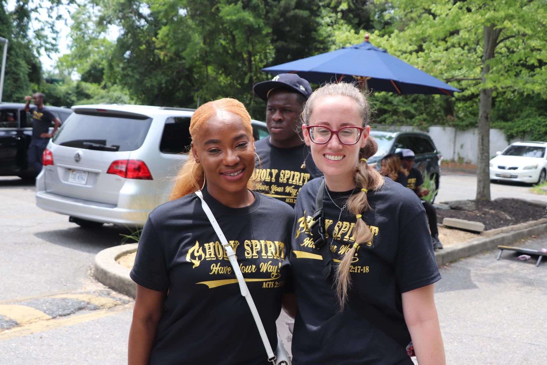 Two women smiling, posing for a photo outside. Both wearing black shirts with gold text. Others in the background.