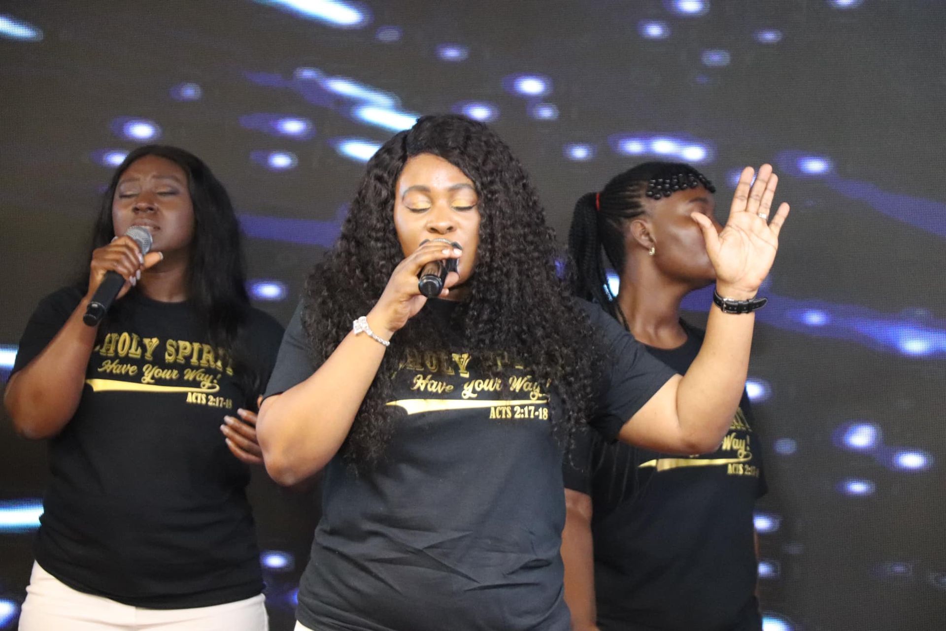 Three women singing on stage, wearing black shirts. Center woman raises hand, singing into mic. Blue background.
