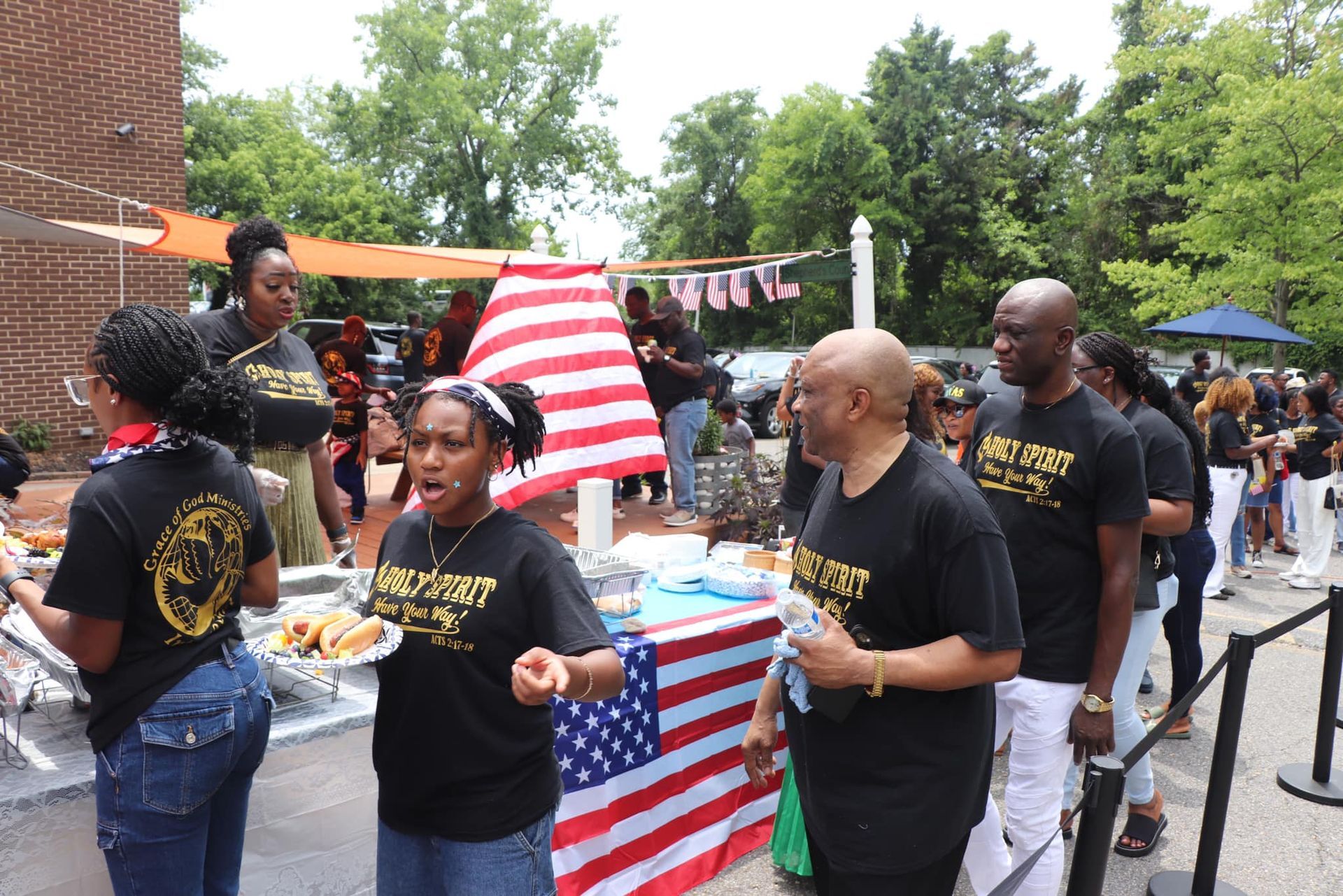 People in black shirts queue for food at an outdoor event with an American flag backdrop.