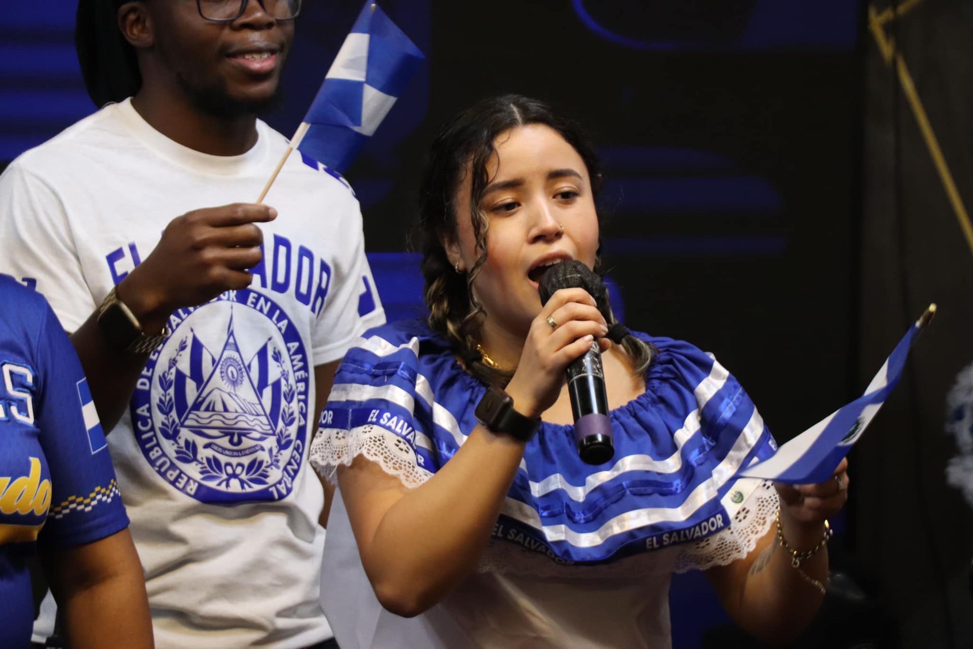 Young woman in blue and white dress singing, holding a Salvadoran flag. Man in background holds flag.