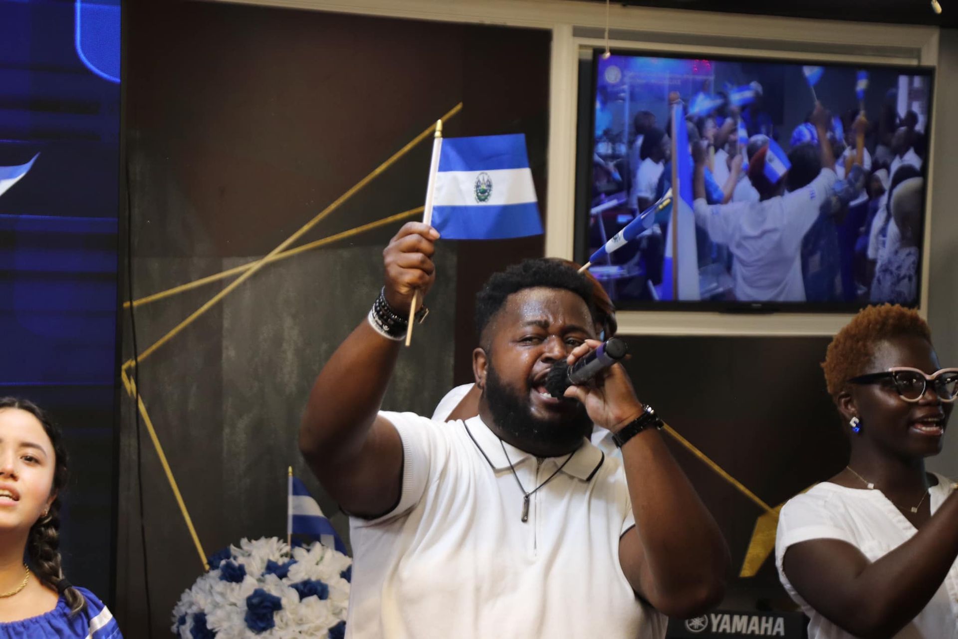 Man singing, waving El Salvador flag. Two women sing along; crowd watches on TV. Blue and white decor.