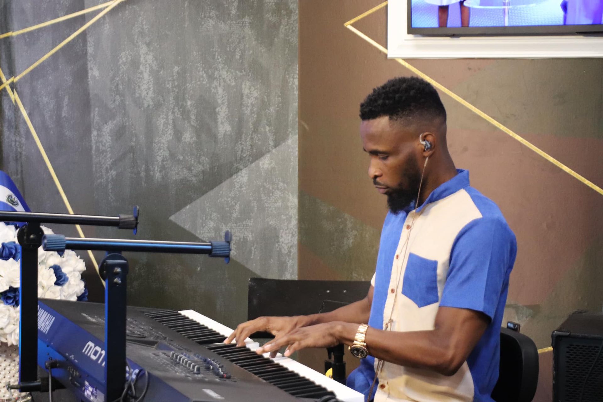 Man playing keyboard onstage, wearing blue and tan shirt. Stage has blue and gold decorations.