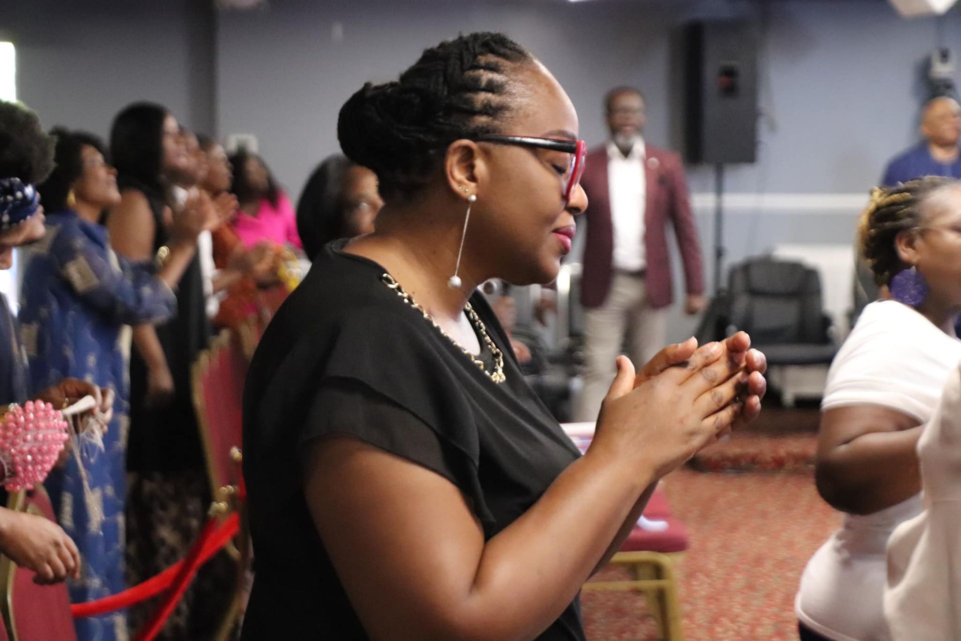 Woman with glasses clapping in a room with others, possibly church.