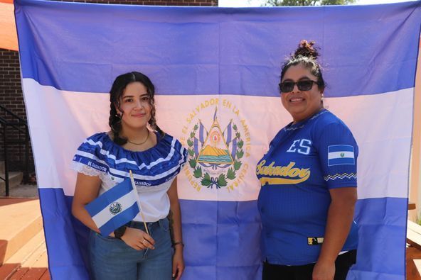 Two women posing in front of the El Salvador flag. One holds a small flag, the other wears a blue shirt.