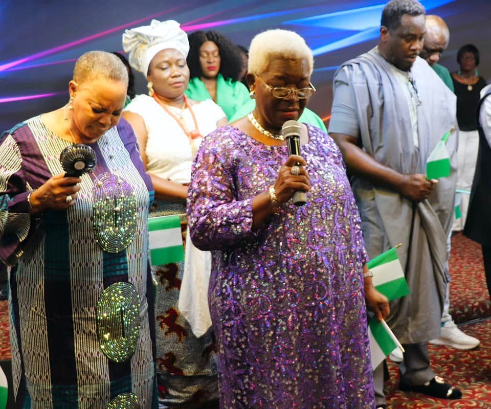 People at a ceremony, woman speaking into a microphone, others holding flags.