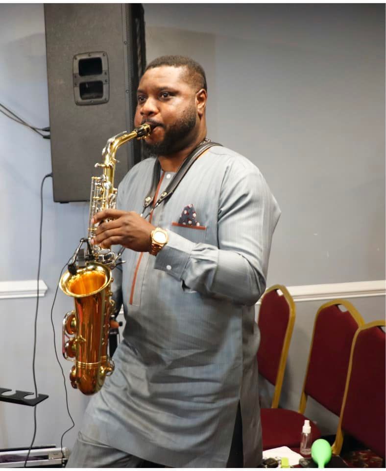 Man playing a gold saxophone, wearing a gray shirt. Indoors, near chairs and a speaker.