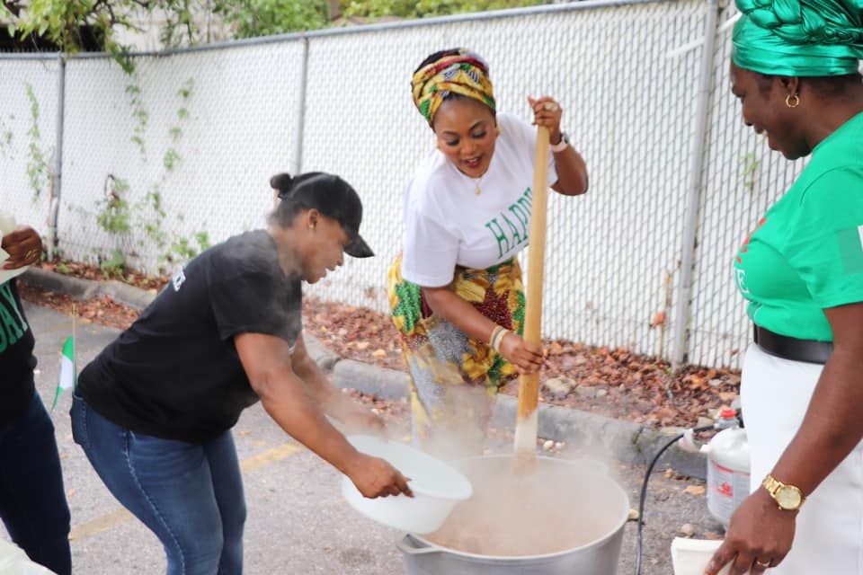 Three women cook outside, stirring a large pot. One adds water, all wearing green and white clothing.