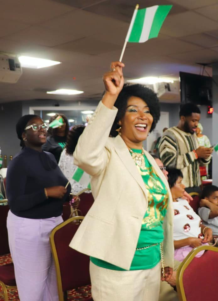 Woman in green and cream, waving a Nigerian flag, smiles enthusiastically in a crowded room.