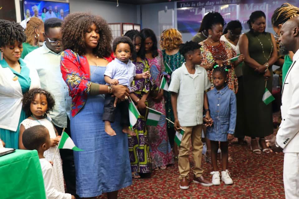People holding Nigerian flags gather in a room, dressed in green and white for a celebration.
