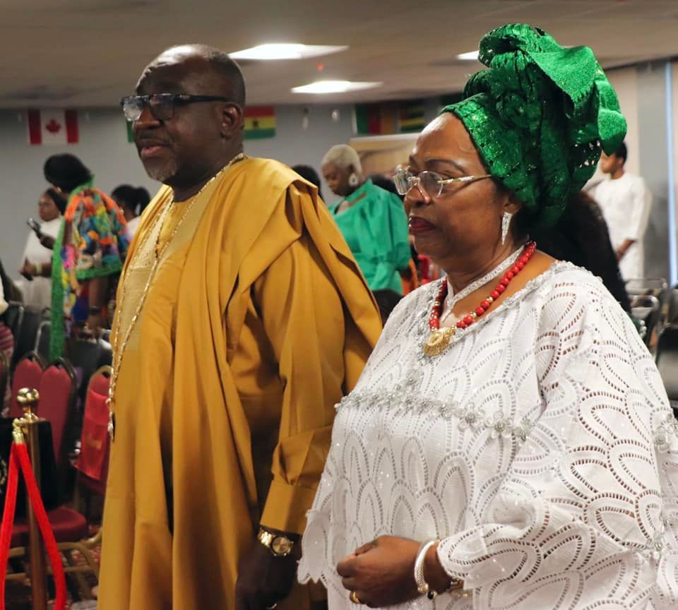 Man and woman in traditional African attire, indoors. Man wears gold, woman wears white and green headwrap.