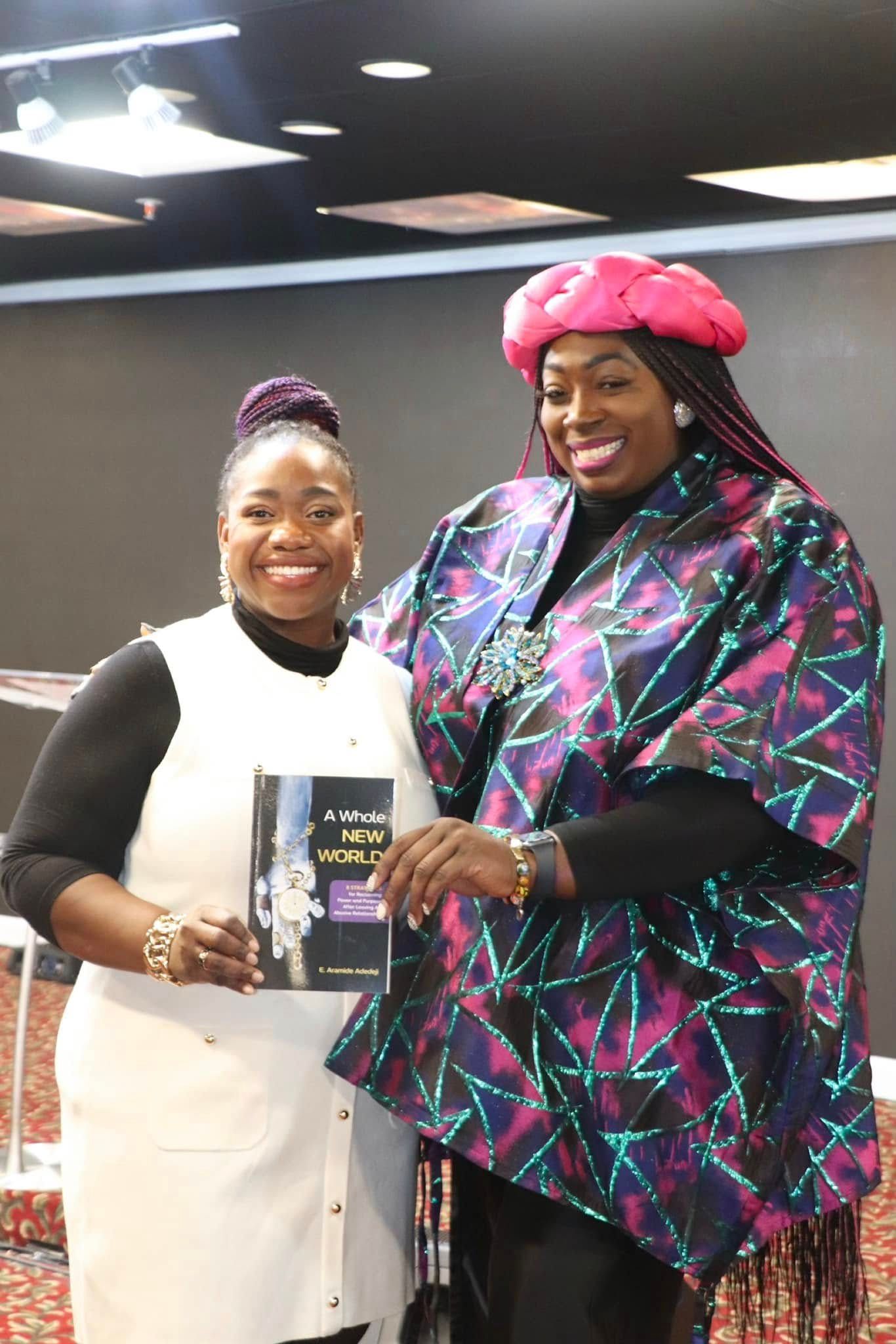 Two women smiling, one holding a book, in a room with a red carpet.