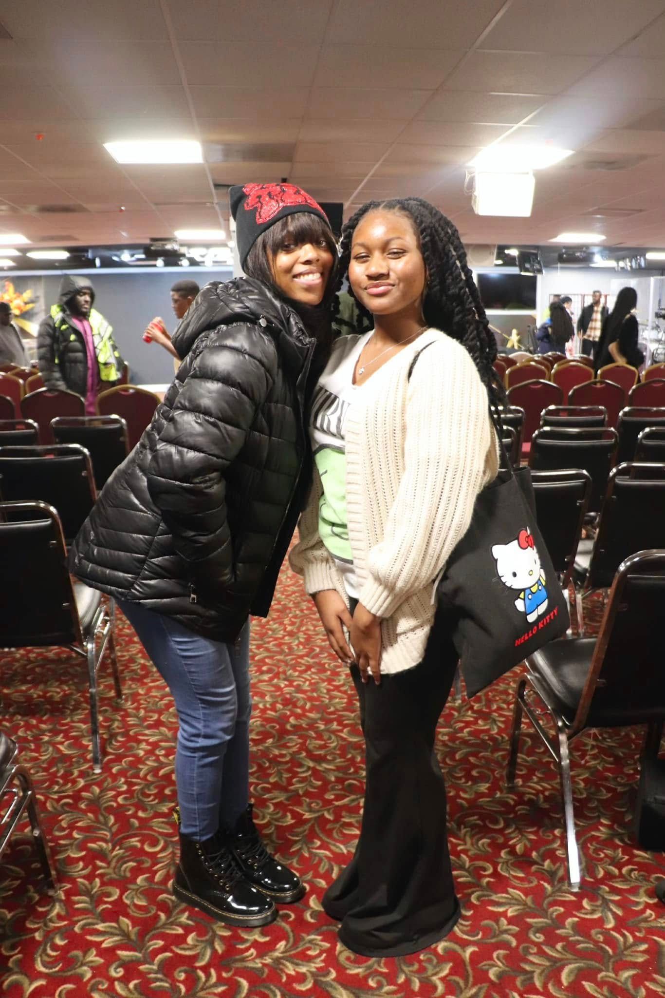 Two women smiling, posing together in a room with chairs, one in a black coat, the other in a sweater.