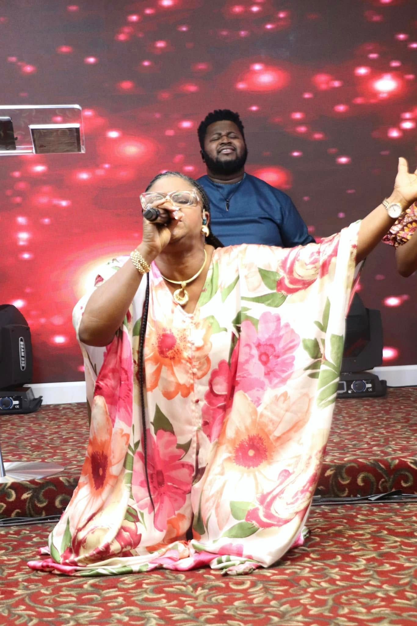 Woman singing on her knees in front of a stage. Man in the background. Red and gold backdrop.
