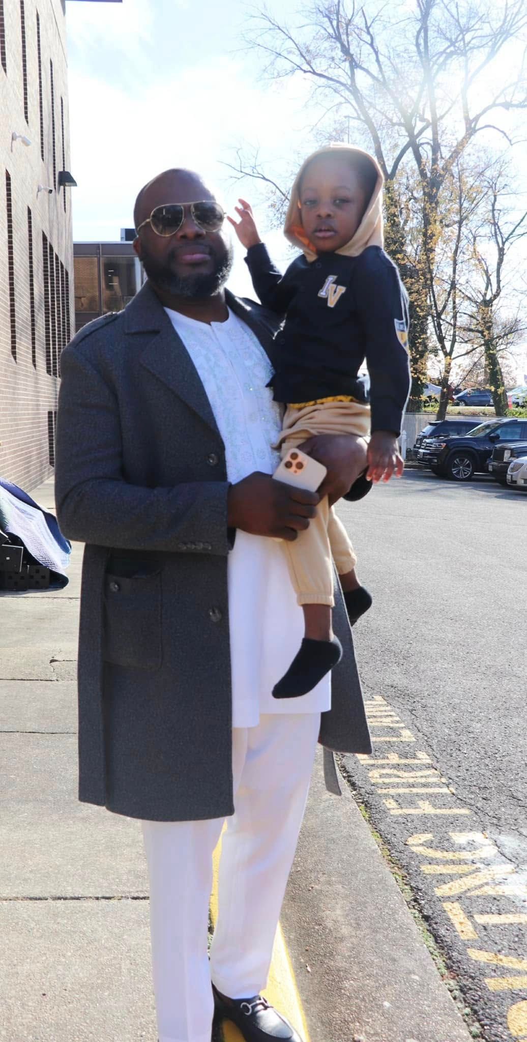 Man in gray coat holding a child. They stand on a city street. Sunny day.