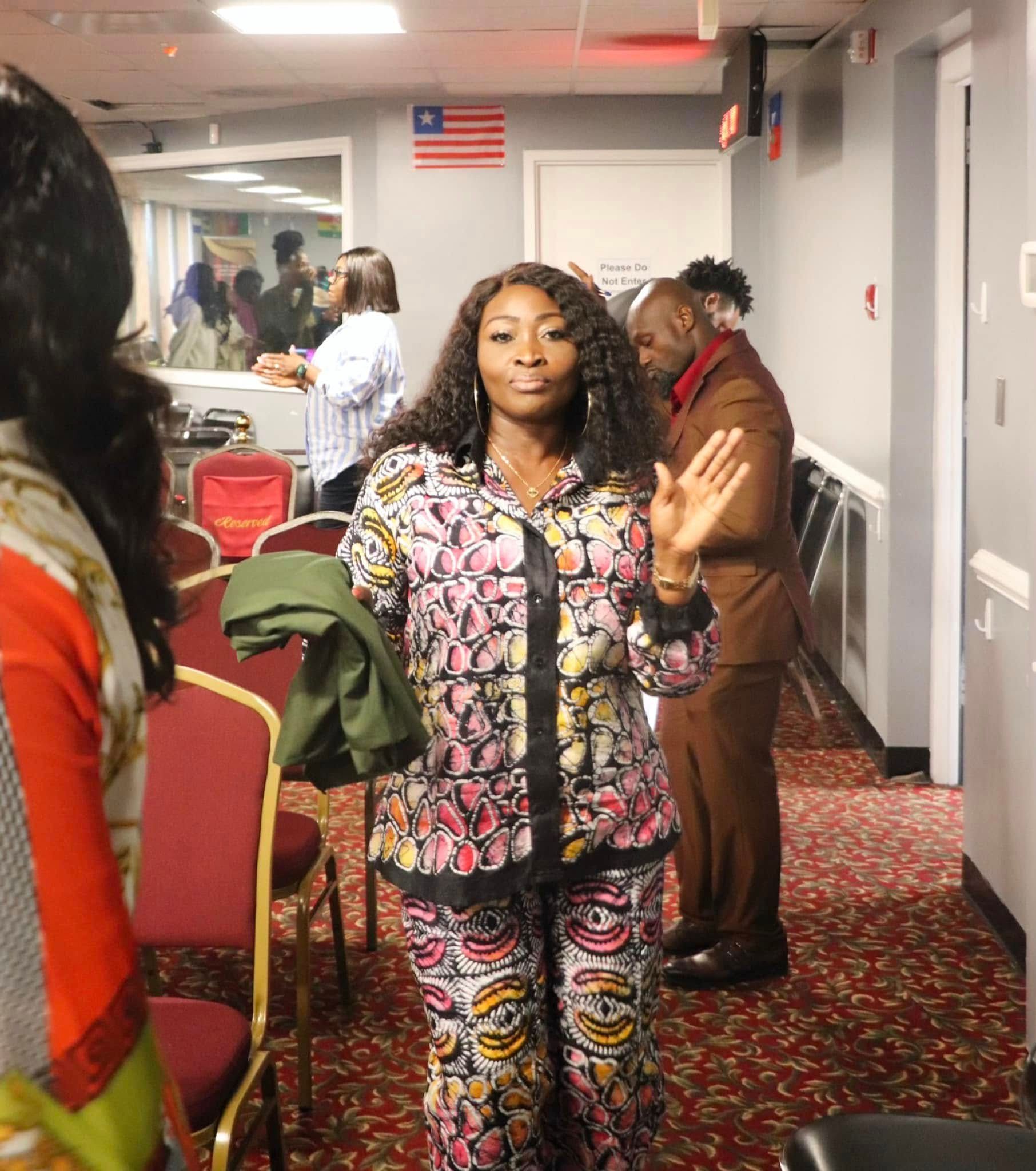 Woman in patterned outfit raises hand, standing in a room with others; Liberian flag visible.