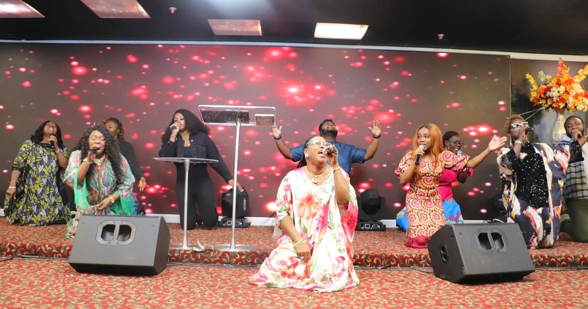 Choir singing on stage with a red and gold backdrop; some raise hands in worship.