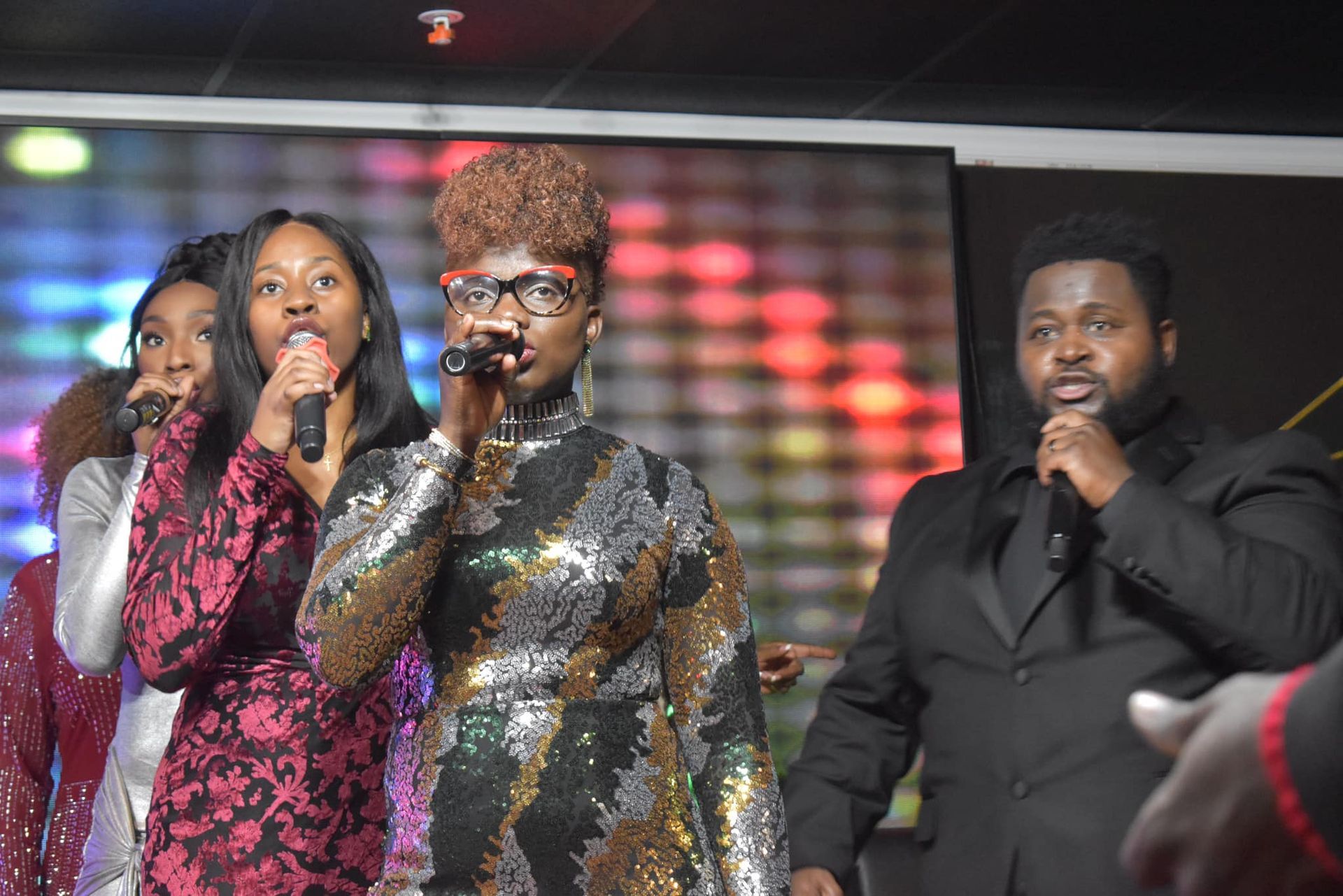 A group of singers on stage, one woman in sparkly dress, others in formal wear, singing into microphones.