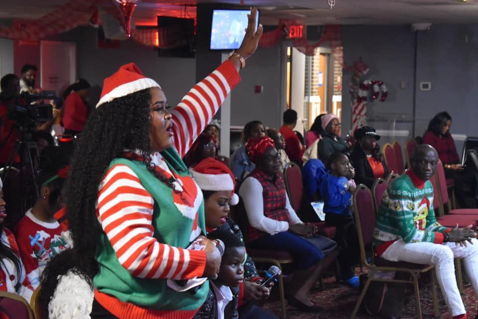 Woman in elf sweater raises arm, singing with a crowd in Christmas sweaters.