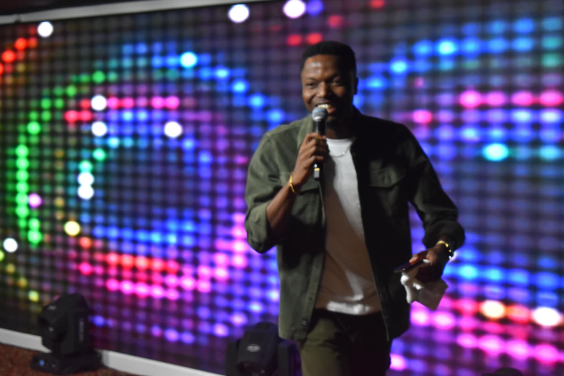 Man on stage with microphone, speaking. Colorful background.
