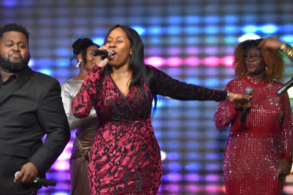 A gospel singer in a red dress performs onstage, surrounded by backup singers against a colorful background.