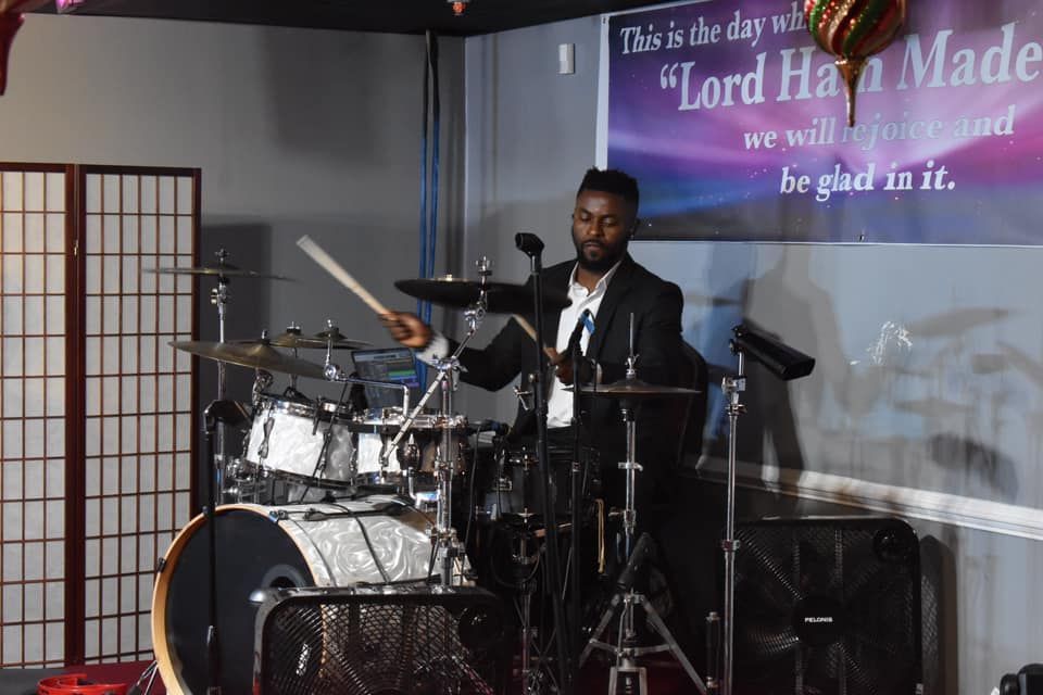 Drummer in a suit playing a drum kit indoors, with a decorative screen and banner.