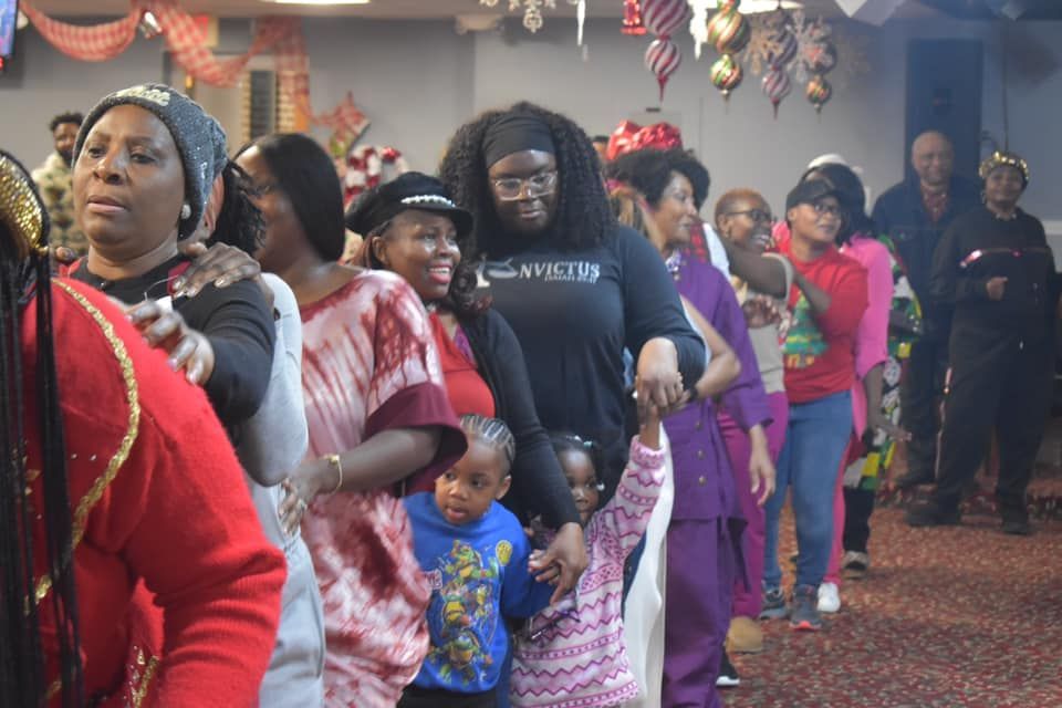 People of different ages dancing in a line at a gathering. Festive decorations in the background.