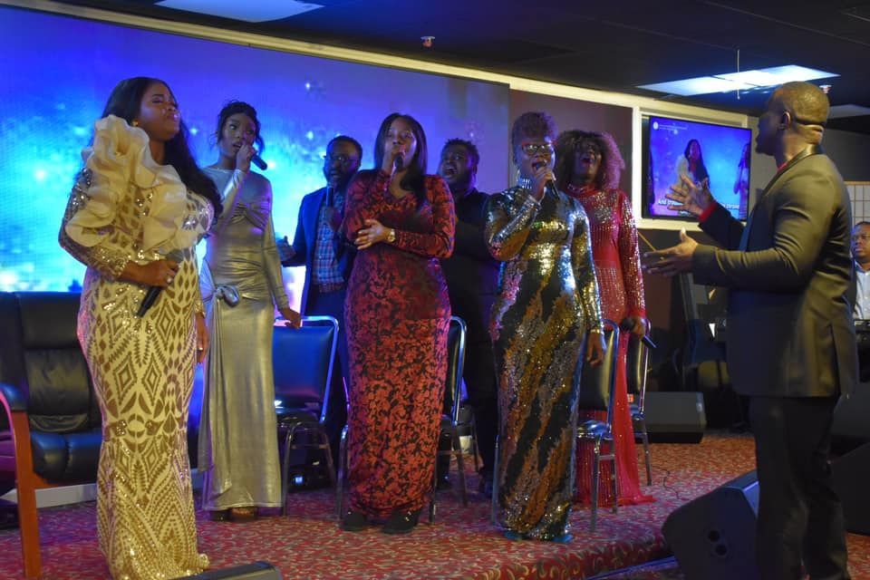 Choir singing on stage, several women in gowns, man conducting. Blue background, red carpet stage.