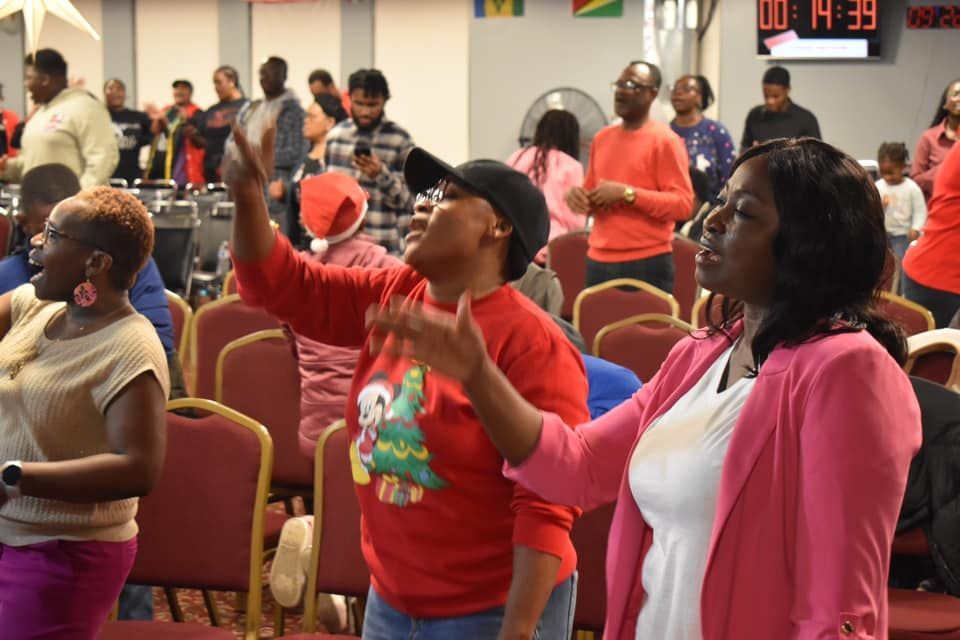 People in a church service, many with hands raised, worshipping. Red and pink clothing, indoors.