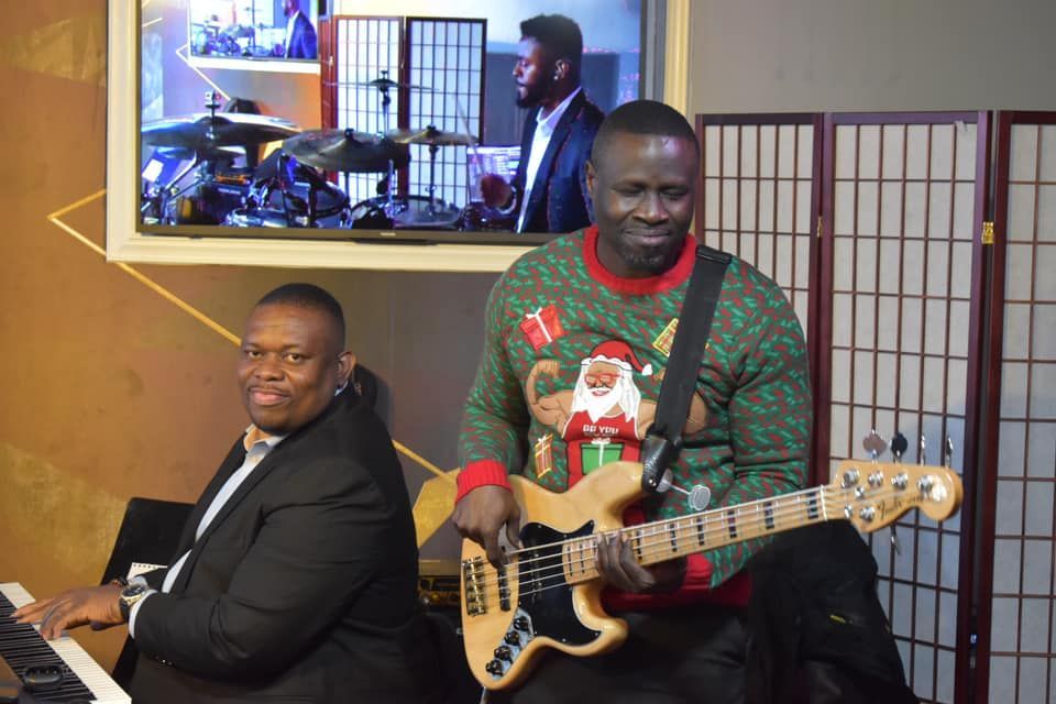 Two Black men playing music in a studio: a pianist and a bassist wearing a Christmas sweater. A screen displays a band.