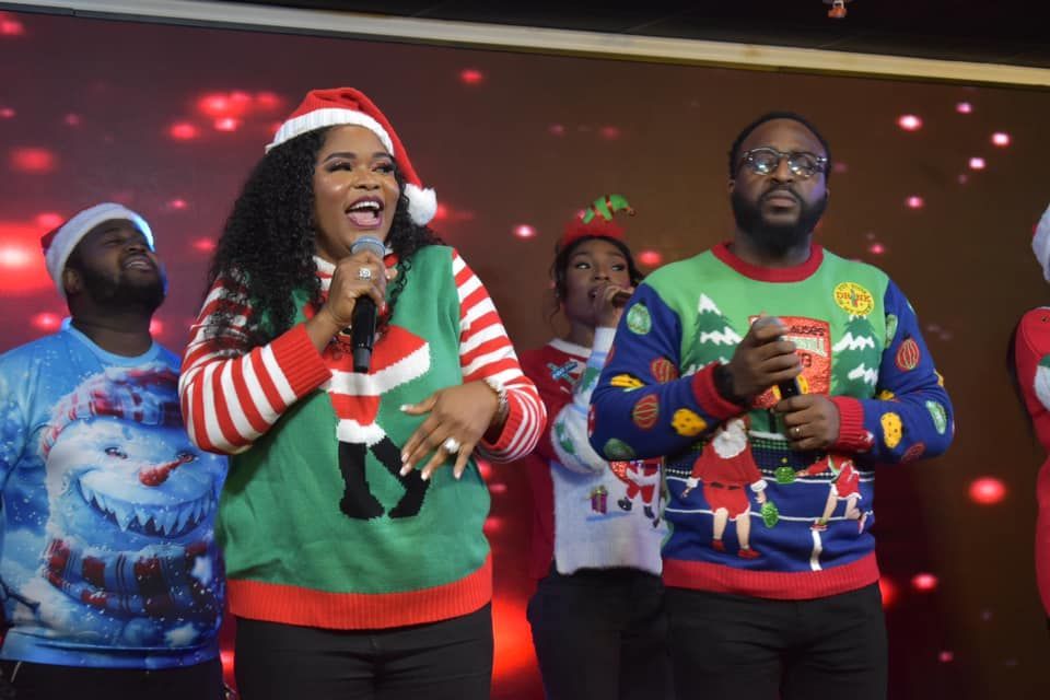 Choir singing Christmas carols onstage, wearing festive sweaters and hats. Red and white decorations.