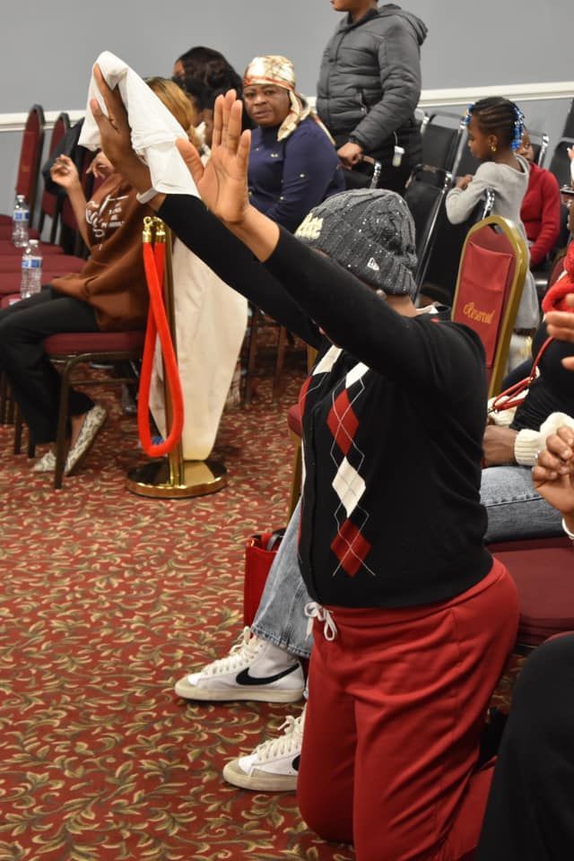 Woman kneels, arms raised in prayer. Red sweatsuit, black sweater with argyle. Church interior.