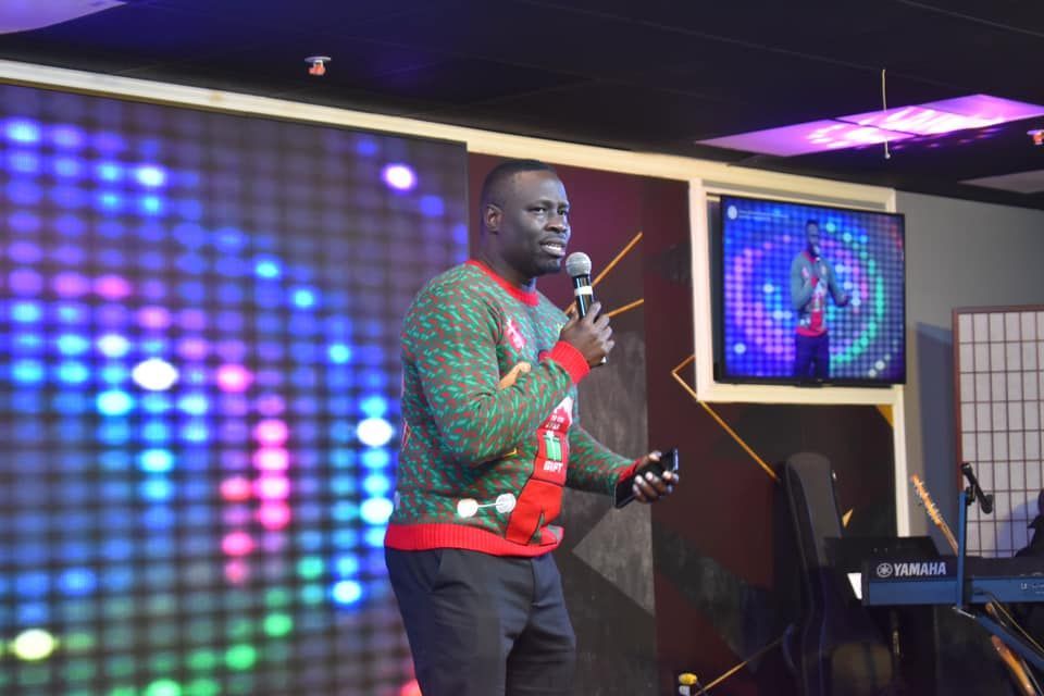 Man in Christmas sweater speaks into a microphone on stage with a colorful LED backdrop and a TV screen displaying him.