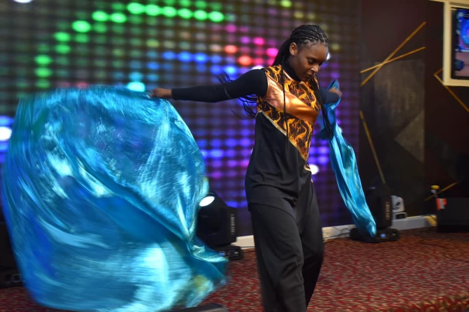 Person in black outfit with gold accents twirls bright blue flags in front of a colorful screen.