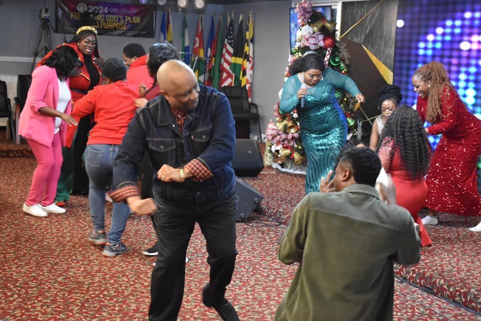 Group of people dancing on stage, a church setting. Some wear red outfits, joyful expressions.