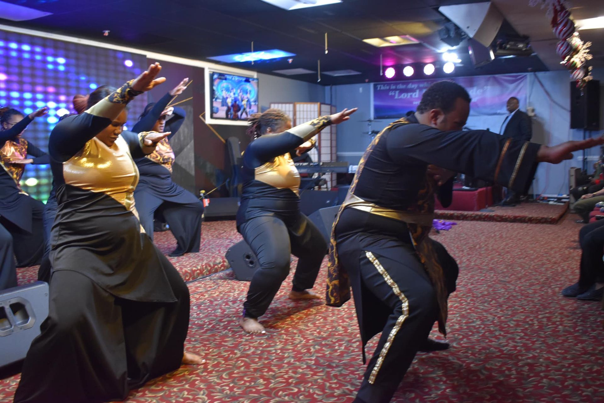 Group of people dancing on stage with arms raised, wearing black and gold outfits; a brightly lit indoor setting.