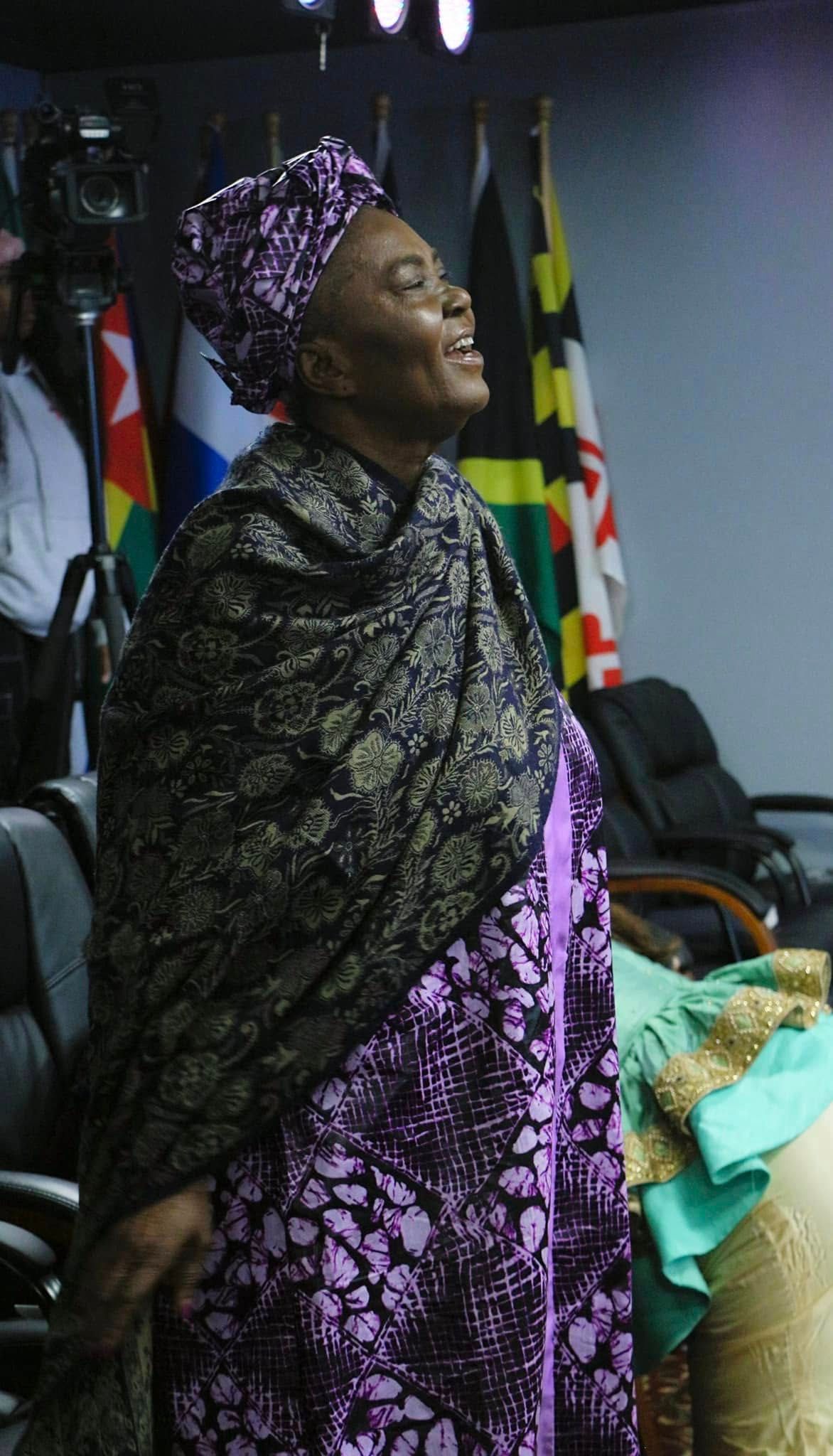 Woman in purple and black patterned attire with head wrap, smiling upwards, indoors. Flags in background.