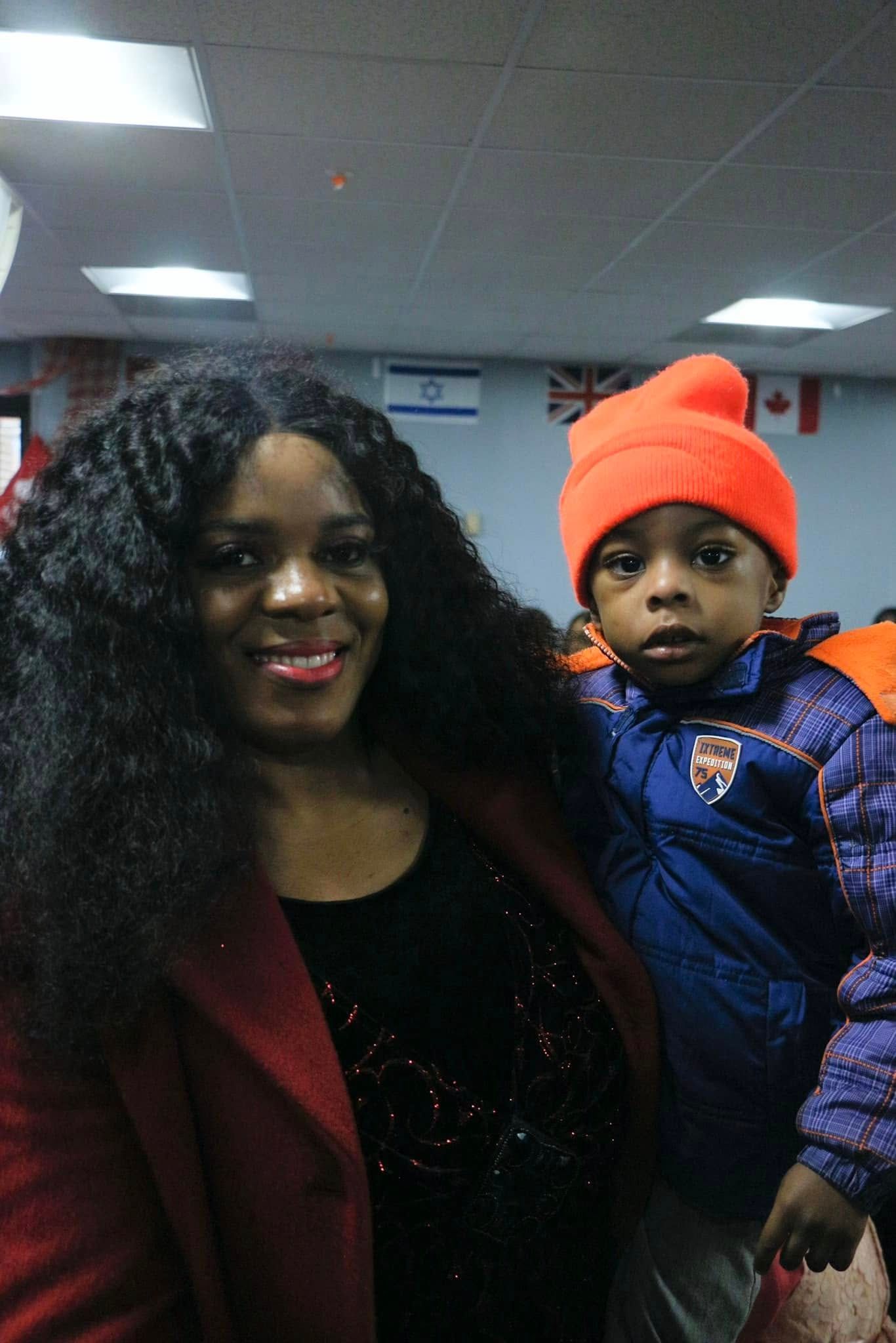 Woman with long dark hair smiles, holding a child wearing an orange hat and blue jacket. Indoor setting.