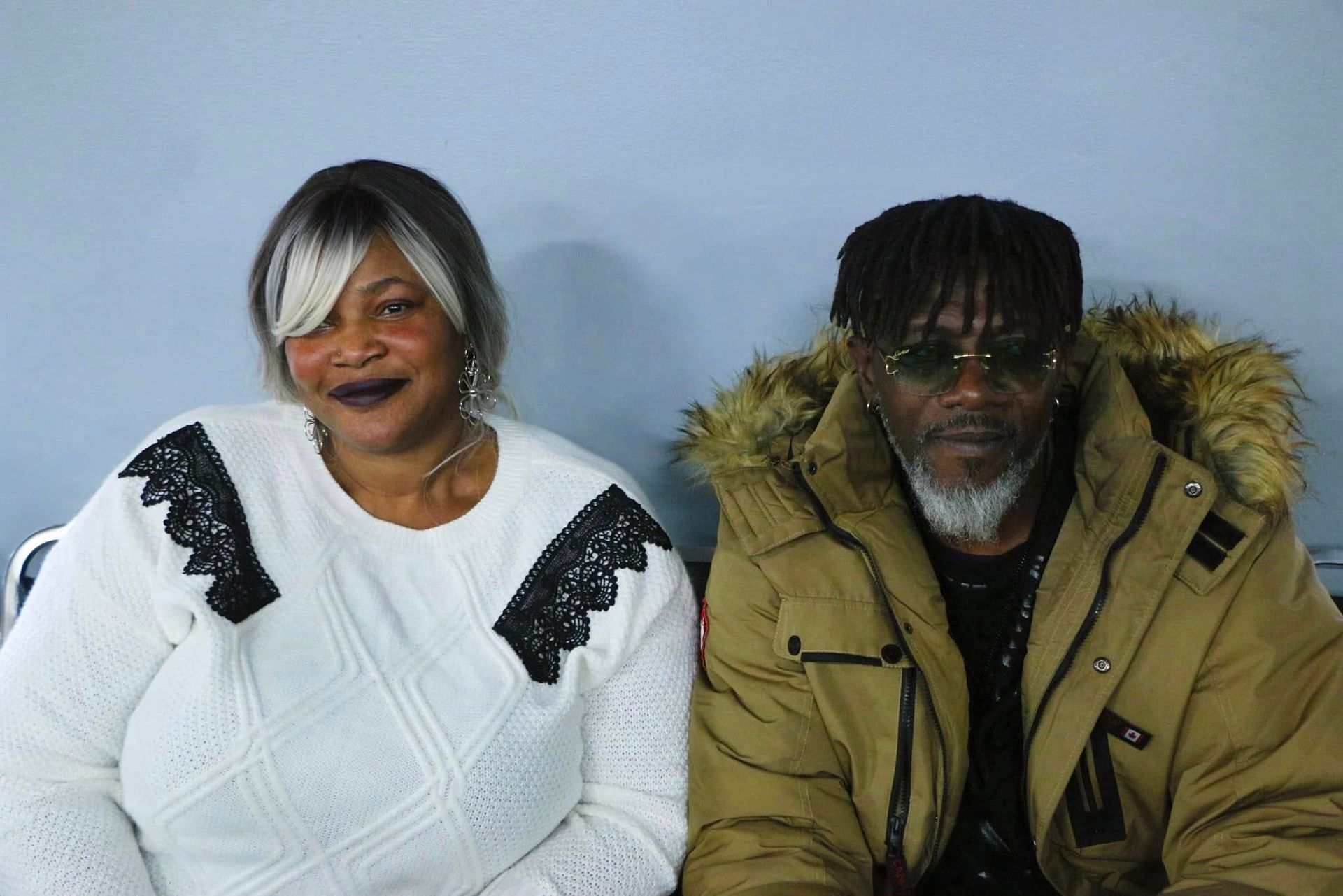 Woman with blonde and black hair sits next to a man in a brown jacket, they are indoors.