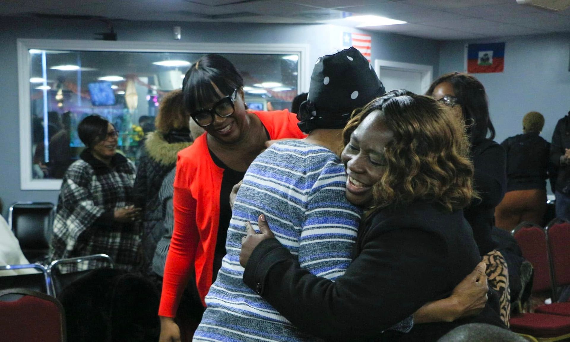 Three women embracing joyfully indoors, smiling, with a Haitian flag visible in the background.
