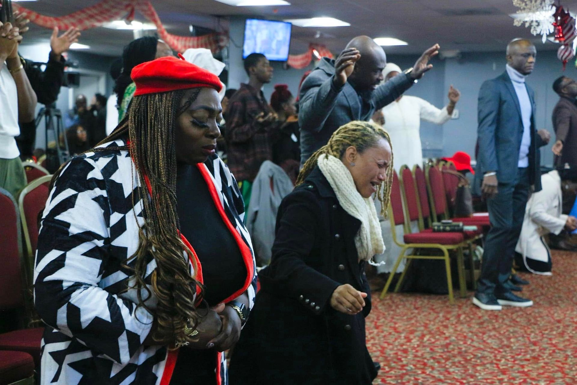 People praying in a church; some with arms raised, others with heads bowed. Red, black, and white clothing.