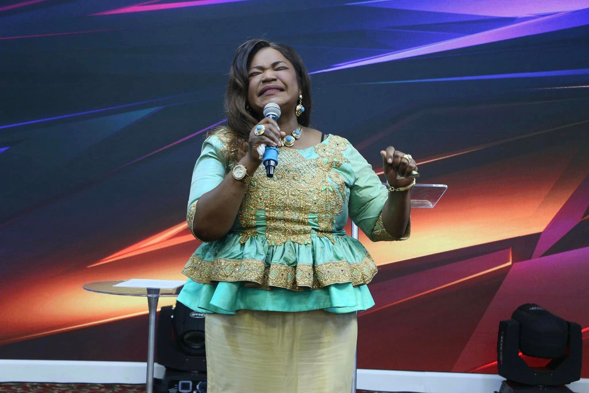 Woman singing into a microphone on a stage, wearing a teal and gold outfit.