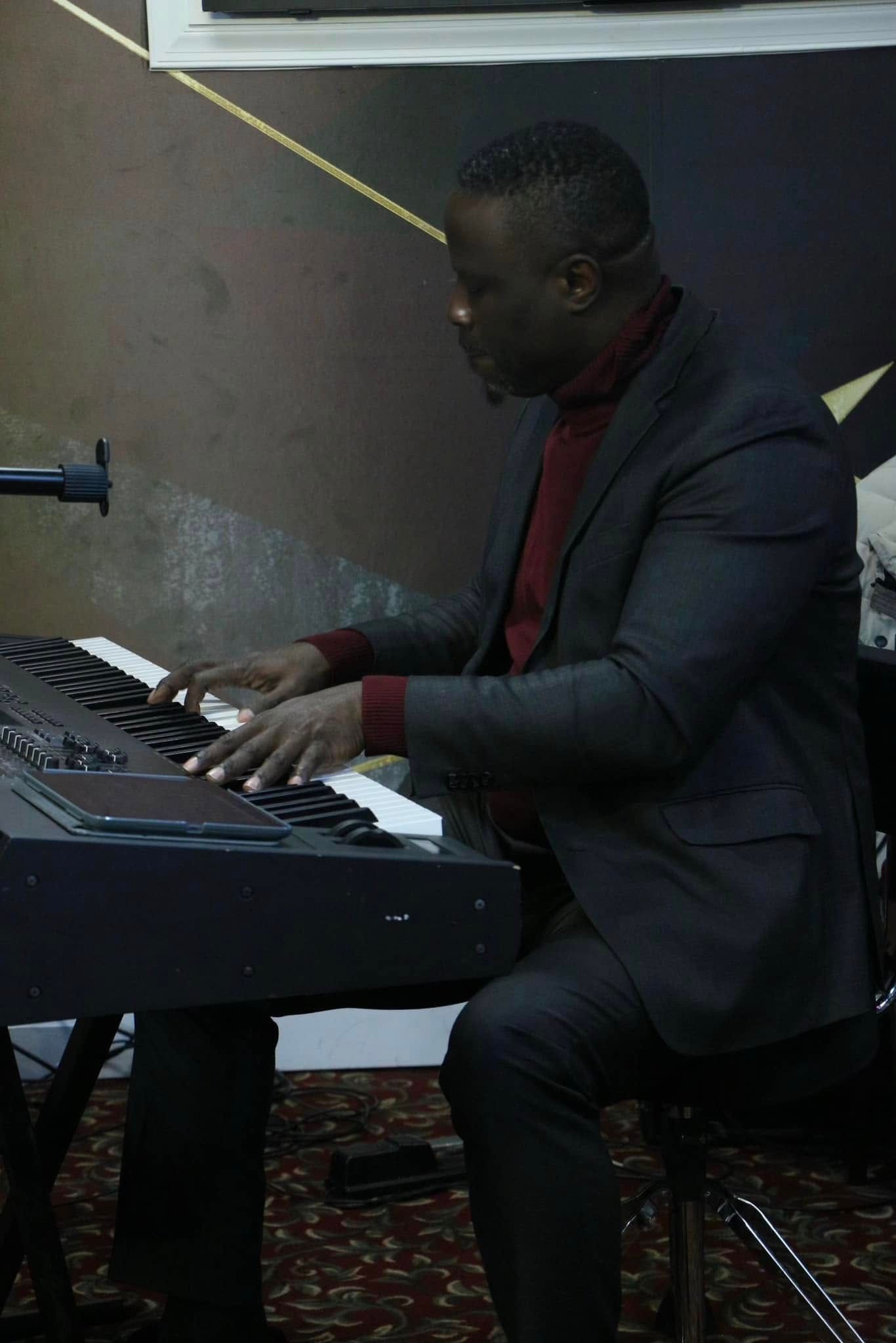 Man playing a keyboard, wearing a dark suit and maroon sweater. Indoor setting with a microphone.